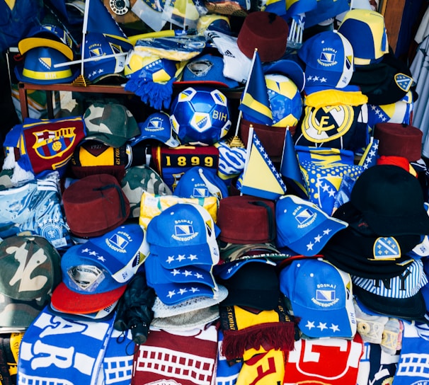 A vibrant display of sports merchandise with various hats, caps, and flags in blue and yellow, prominently featuring the Bosnia and Herzegovina flag and football-related items. There are shirts and scarves with team logos, including a jersey with the name Mbappe and the number 29.
