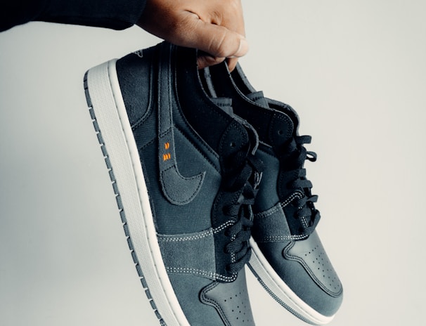 A hand is holding a pair of dark-colored athletic shoes. The shoes have a combination of leather and mesh materials with a swoosh logo on the side. They feature black laces and white soles. The hand appears to be wearing a black sleeve.